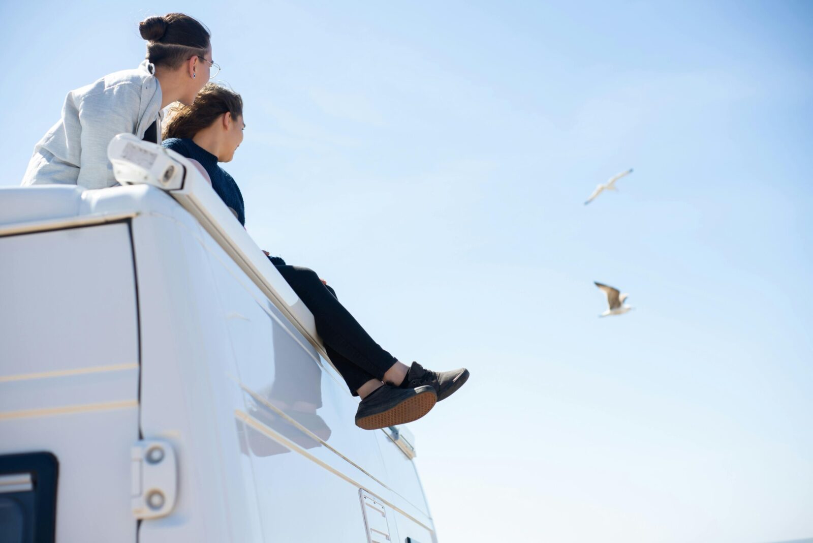 Two people sitting on a camper van roof enjoying a sunny day with seagulls flying.